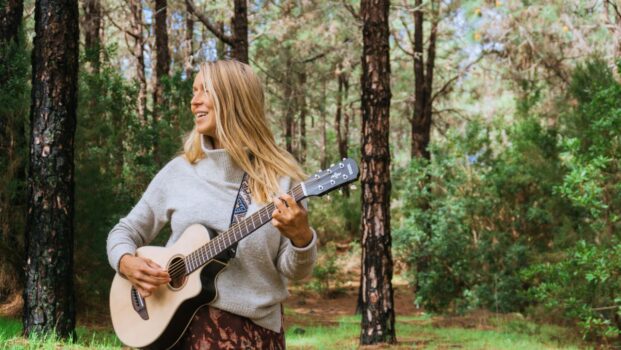Vaaleahiuksinen nainen seisoo aurinkoisessa metsässä ja soittaa kitaraa.