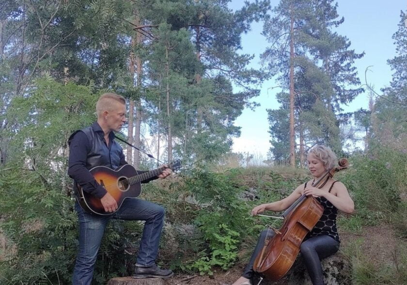 Duo musisoi metsäisessä maisemassa. Sellisti istuu kallion reunalla ja kitaristi seisoo häntä vastapäältä.