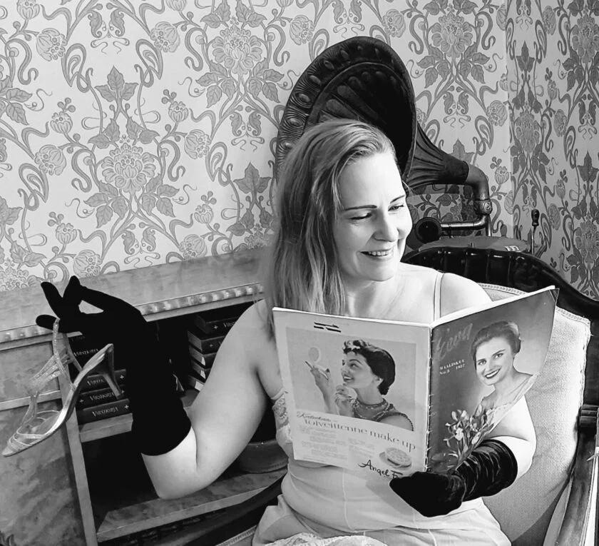Mustavalkoinen kuva. Kuvassa istuva nainen katsoo lehteä, jonka kansissa on kuvia naisista. Nainen hymyilee. Naisen kädessä hansikkaat ja sormesta roikkuva korkokenkä. Taustalla vanha gramofoni ja tapetoitu seinä.