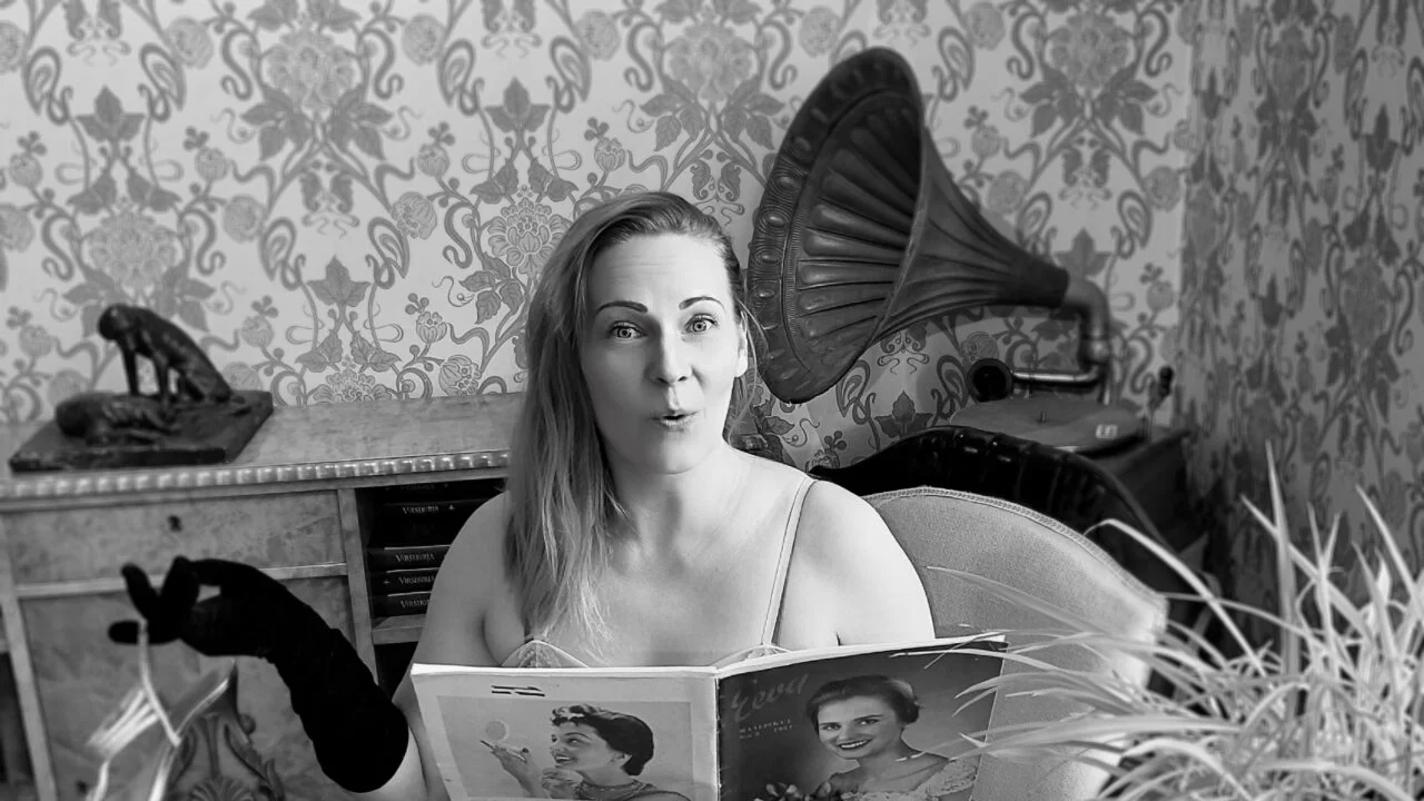 Mustavalkoinen kuva. Kuvassa istuva nainen lukee lehteä, jonka kansissa on kuvia naisista. Nainen katsoo kohti kameraa. Naisen kädessä hansikkaat ja sormesta roikkuva korkokenkä. Taustalla vanha gramofoni ja tapetoitu seinä.