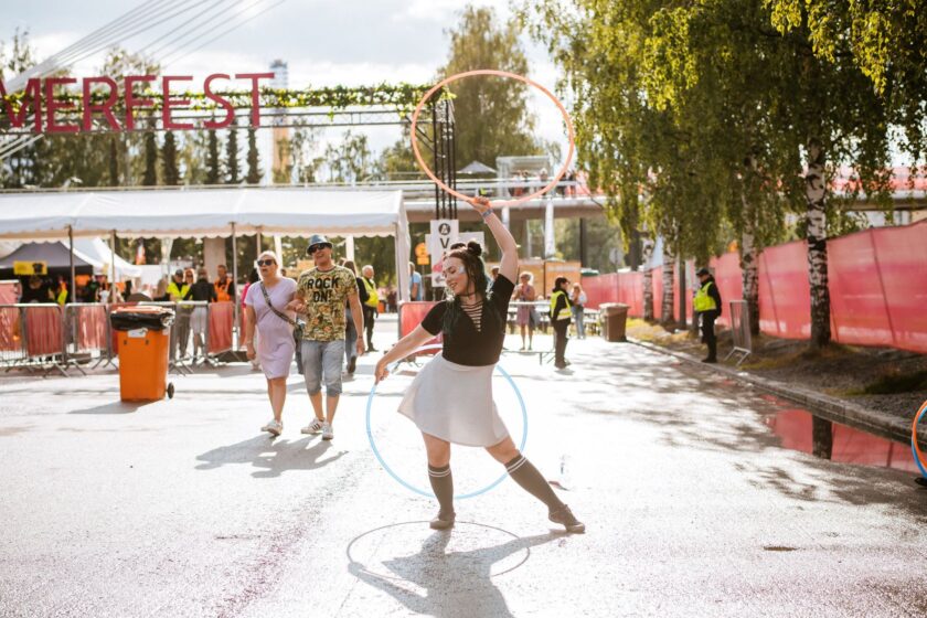 Tammerfestien portit näkyvät taustalla, festariyleisöä kävelee. Esiintyjä poseeraa kahden vanteen kanssa.