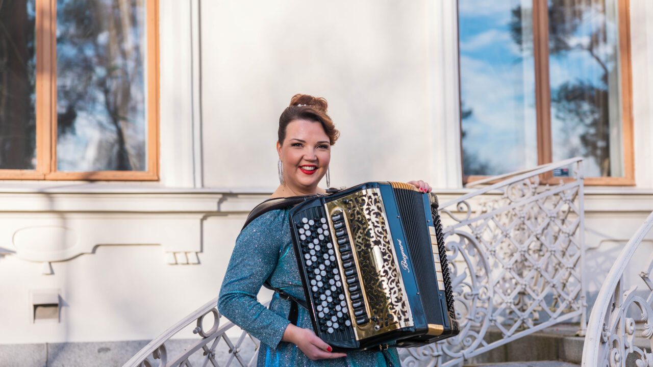 Henna Leppänen seisoo ulkona portailla harmonikka sylissään, kimalteleva iltapuku yllään.