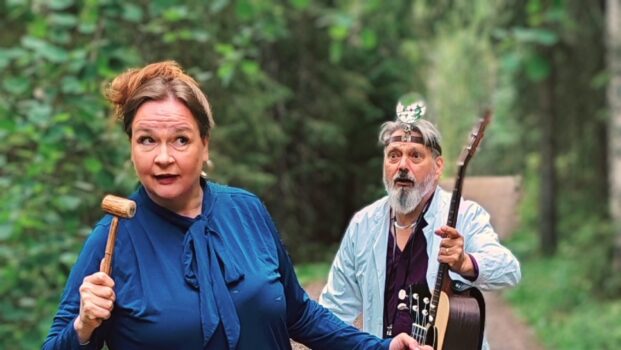 Nainen ja mies kävelevät polulla, naisella kädessään puheenjohtajan vasara ja ukulele. Miehellä lääkärin varusteet ja kitara kädessä.