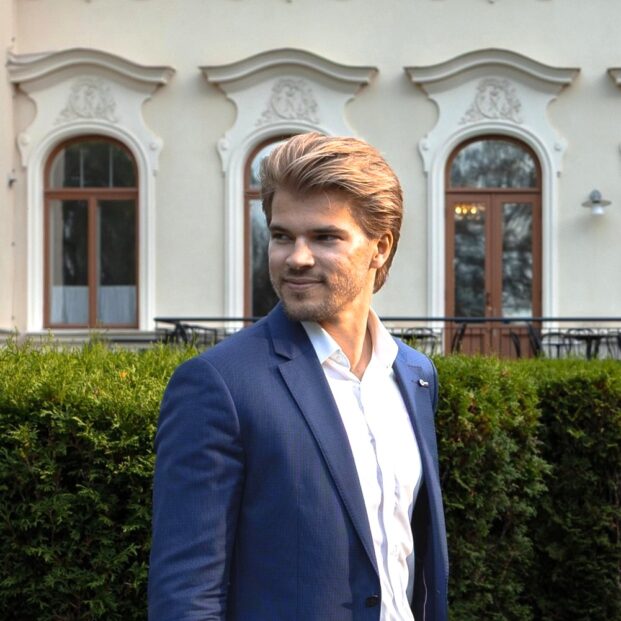 Mies sinisessä puvussa katsoo ohi kameran, taustalla kartano ja puska.