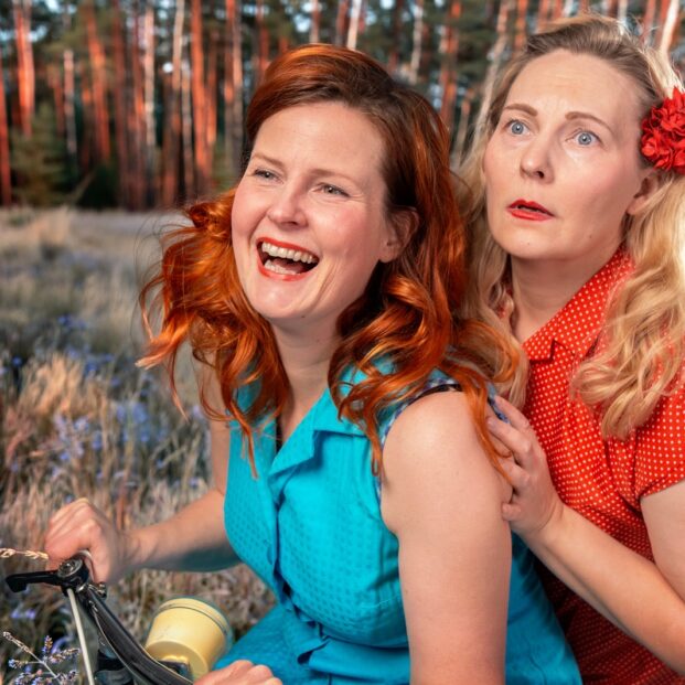 Kaksi naista polkevat kuntopyörän selässä. maalaismaisemassa. Toisella turkoosi mekko, punaiset hiukset ja hymy. Toisella punainen mekko, vaaleat hiukset ja pelokas ilme.