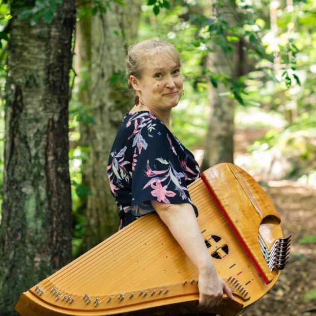Kuvassa nainen seisoo metsäpolulla konserttikantele sylissä.