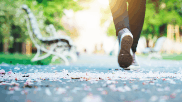Nainen kävelee puiston polkua kohti auringon valoa. Vasemmalla tien vieressä vanha penkki.