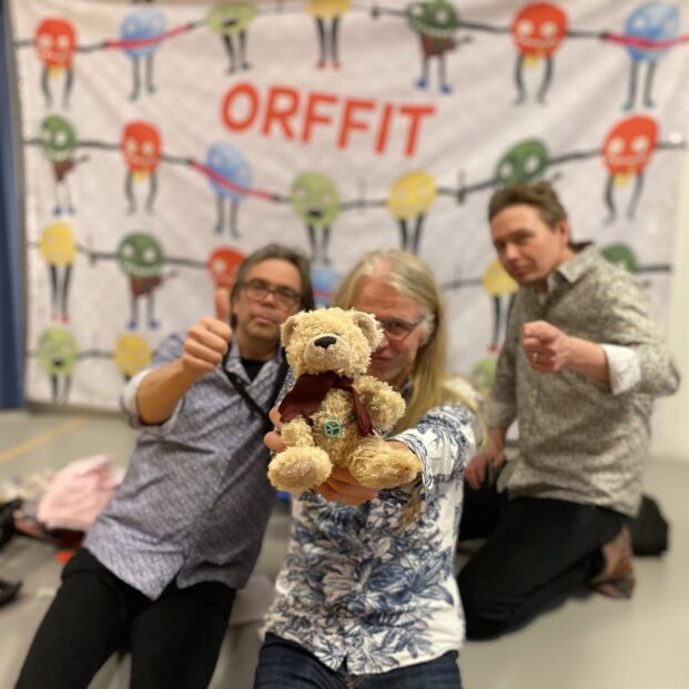 Kuvassa Orffien kolmikko. Vasemmella peukuttaa Matti, oikealla Kimmo. Keskellä kesyttelee hymyään Hannu, jolla on kädessään pieni pehmonalle. Oikeasti nalle on Orffien päällikkö ja vieraileva rumpali. Taustalla Eskari-Ainon piirtämä taideteos Pääjalkaiset. Kuvasta sattaa välittyä innokkaanlempeä, mutta puolivilli suhtaututuminen lastenmusiikin tekemiseen. Jos näin on - se on kolmikon mielestä oikein.