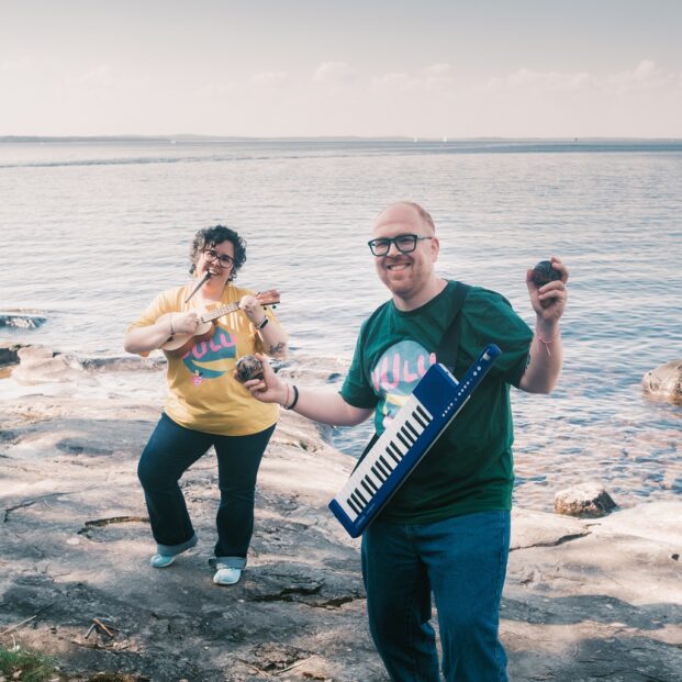 Kuvassa muusikot Paloma Catalá Parga ja Iago Castro Pérez seisovat kallioisella rannalla. Parga seisoo taka-alalla ja soittaa ukulelea pilli suussaan. Etualalla Pérez soittaa marakasseja, ja hänen kaulassaan roikkuu kosketinsoitin olkahihnassa.