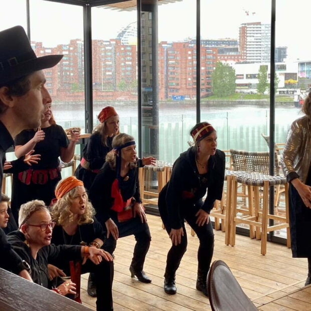 Kuvassa on Tuomiokuoron esiintyjiä sisätilassa, jonka lasiseinän läpi näkyy Tampereen Ratinan maisema. Kuva on otettu esiintymistilanteessa, ja esiintyjät laulavat parhaillaan hieman kyyristyneinä, osa heistä kyykyssä. Kuvan oikeassa reunassa on kuoronjohtaja Petra Poutanen, joka on ottamassa askeleen kohotetun tason päälle.