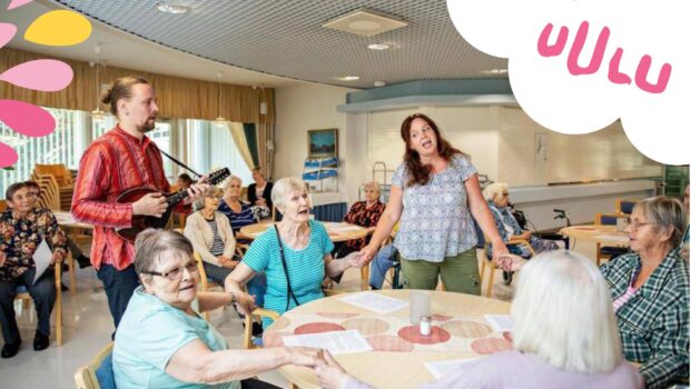 Kuvassa muusikot soittavat ja laulavat ikäihmisten palvelukodin salissa yhdessä asukkaiden kanssa. Keskellä on pyöreä pöytä, jonka ympärillä istuvat henkilöt pitävät toisiaan käsistä kiinni muodostaen laulavan piirin. Vasemmassa reunassa on kitaransoittaja ja taaempana lisää laulavia asukkaita. Kuva on värikäs ja oikeassa yläreunassa on valkoisen pilven päällä Uulun vaaleanpunainen logo.