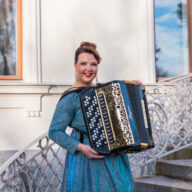 Henna Leppänen seisoo ulkona portailla harmonikka sylissään, kimalteleva iltapuku yllään.