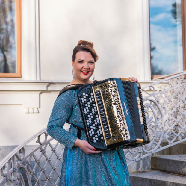 Henna Leppänen seisoo ulkona portailla harmonikka sylissään, kimalteleva iltapuku yllään.