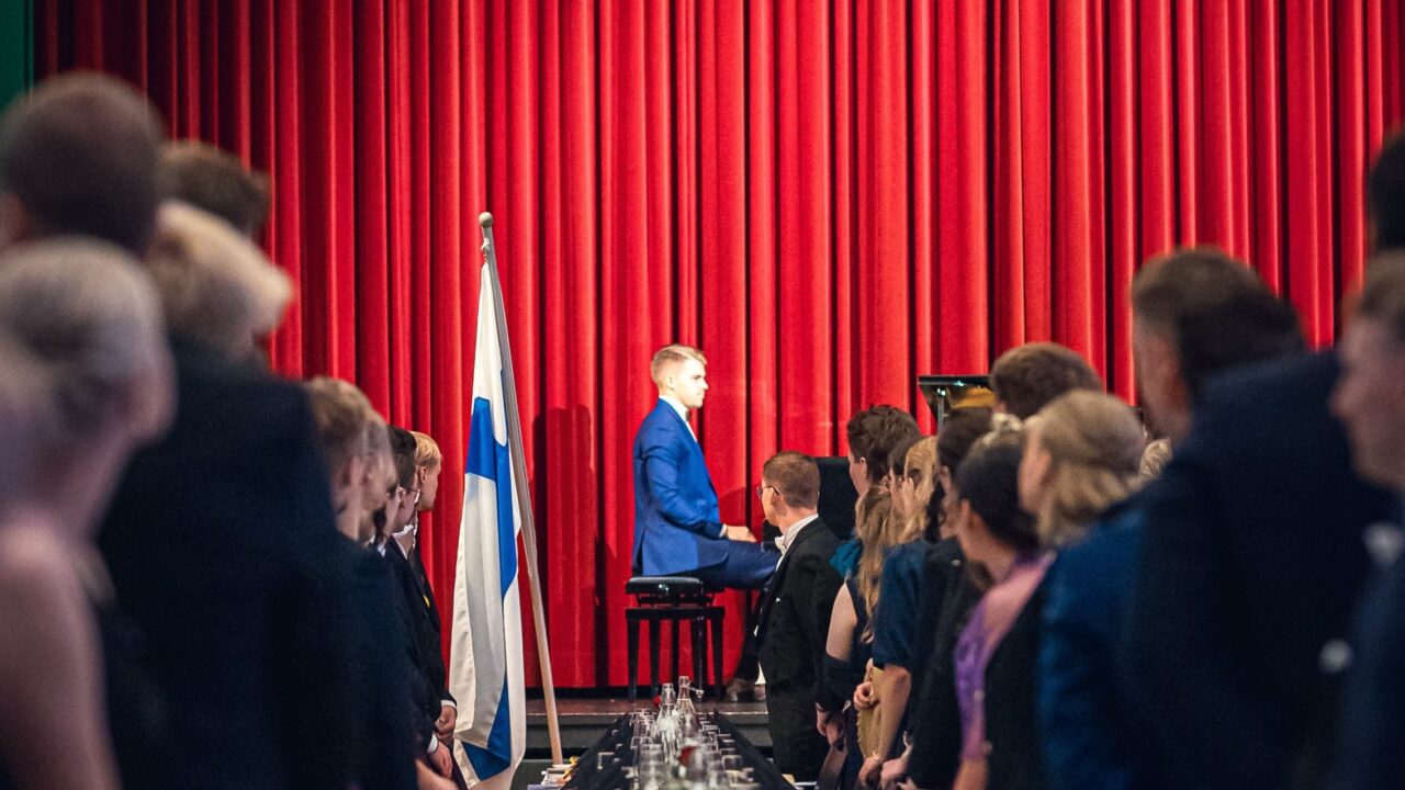 Juhlava juhlasalikuva, jossa pitkän, mustaliinaisen juhlapöydän molemmilla puolilla seisoo juhlavasti pukeutuneita ihmisiä. Kuvan keskellä, lavan edustalla kirkkaanpunaisen teatteriverhon edessä, istuu siniseen pukuun pukeutunut nuori mies pianon ääressä esiintymisvalmiina. Pianistin vieressä seisoo Suomen lippu. Katsojat katsovat kohti pianoa seisten, tunnelma on arvokas ja odottava.
