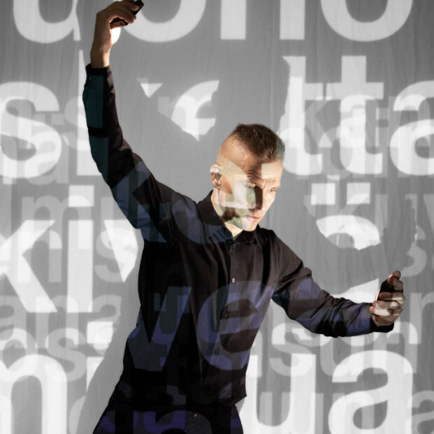 Kuvan keskellä on henkilöllä, jolla on tummat vaatteet ja hänen oikea käsi osoittaa alas ja vasen käsi ylös. Hänen taustalleen on projisoituna tekstiä, joka heijastuu myös henkilön kasvoihin ja vaatteisiin.