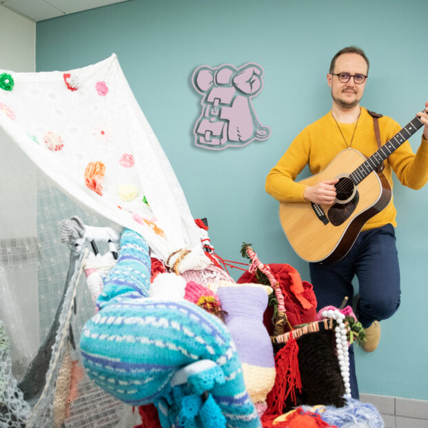 Muusikko soittaa akustista kitaraa huoneessa, jossa on lasten leikkiteltta ja pehmoleluja. Tunnelma on lämmin ja yhteisöllinen.