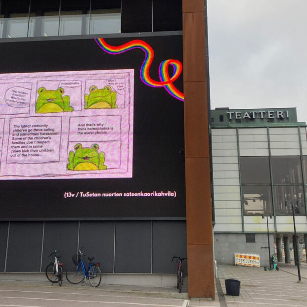 Kuvassa on Turun kaupunginteatterin taideseinä (digitaalinen led-näyttö), johon on heijastettu nuoren tekemä sarjakuva sammakosta. Sarjakuva on englanniksi. Kuvassa on myös pieniä sateenkaarevia nauhoja.