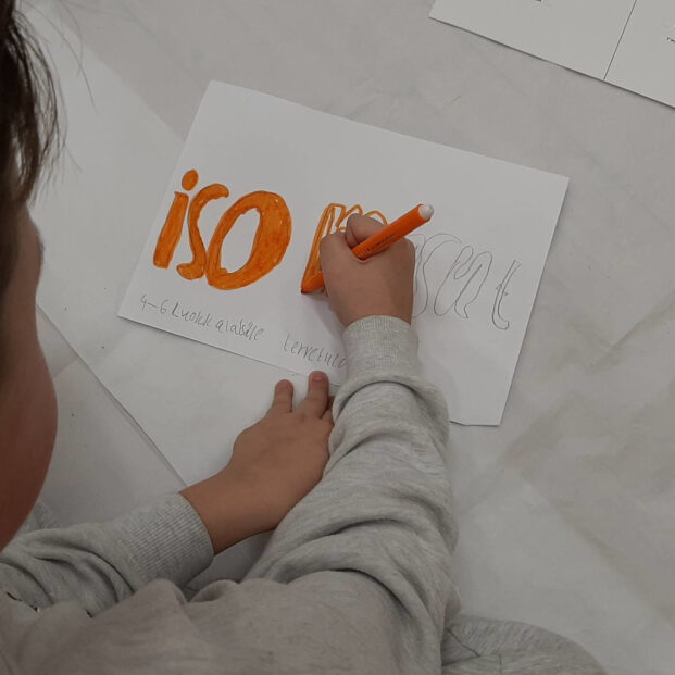 Kuvassa lapsi kirjoittaa lattialla istuessaan paperiin "ISO" oranssilla tussilla.