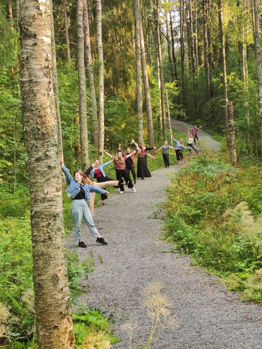 Kuvassa on keväinen metsäpolku, jonka reunassa on nuoria naisia kädet riemukkaasti levällään.