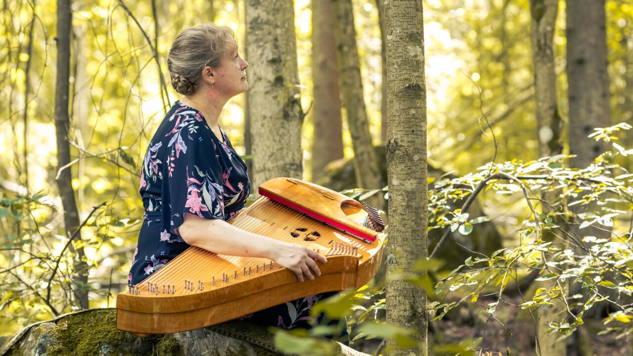 Marja Nevankallio istuu keväisessä metsämaisemassa konserttikantele sylissään, katse suunnattuna sivulle
