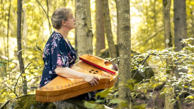 Marja Nevankallio istuu keväisessä metsämaisemassa konserttikantele sylissään, katse suunnattuna sivulle