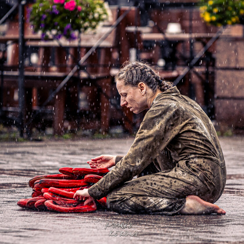 Kuvassa esiintyjä polvistuu ja kiinnittää punaisia kenkiä reppuunsa samalla kun hän itkee sateessa. In the image, a performer is kneeling and attaching red shoes to a backpack while crying in the rain.